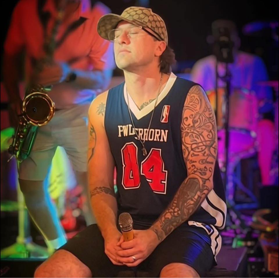 A. Wise performing live, seated with a mic, sax and drums behind him