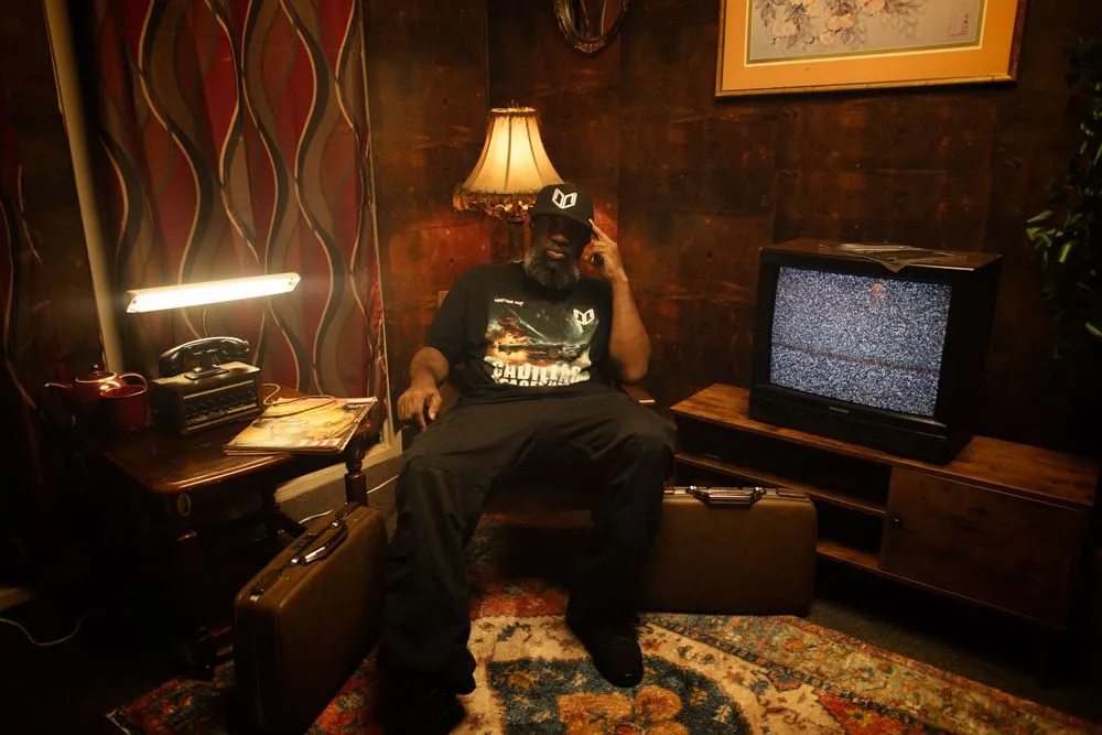 Master Wiz, seated in a vintage wood-paneled room with a CRT TV and lamp